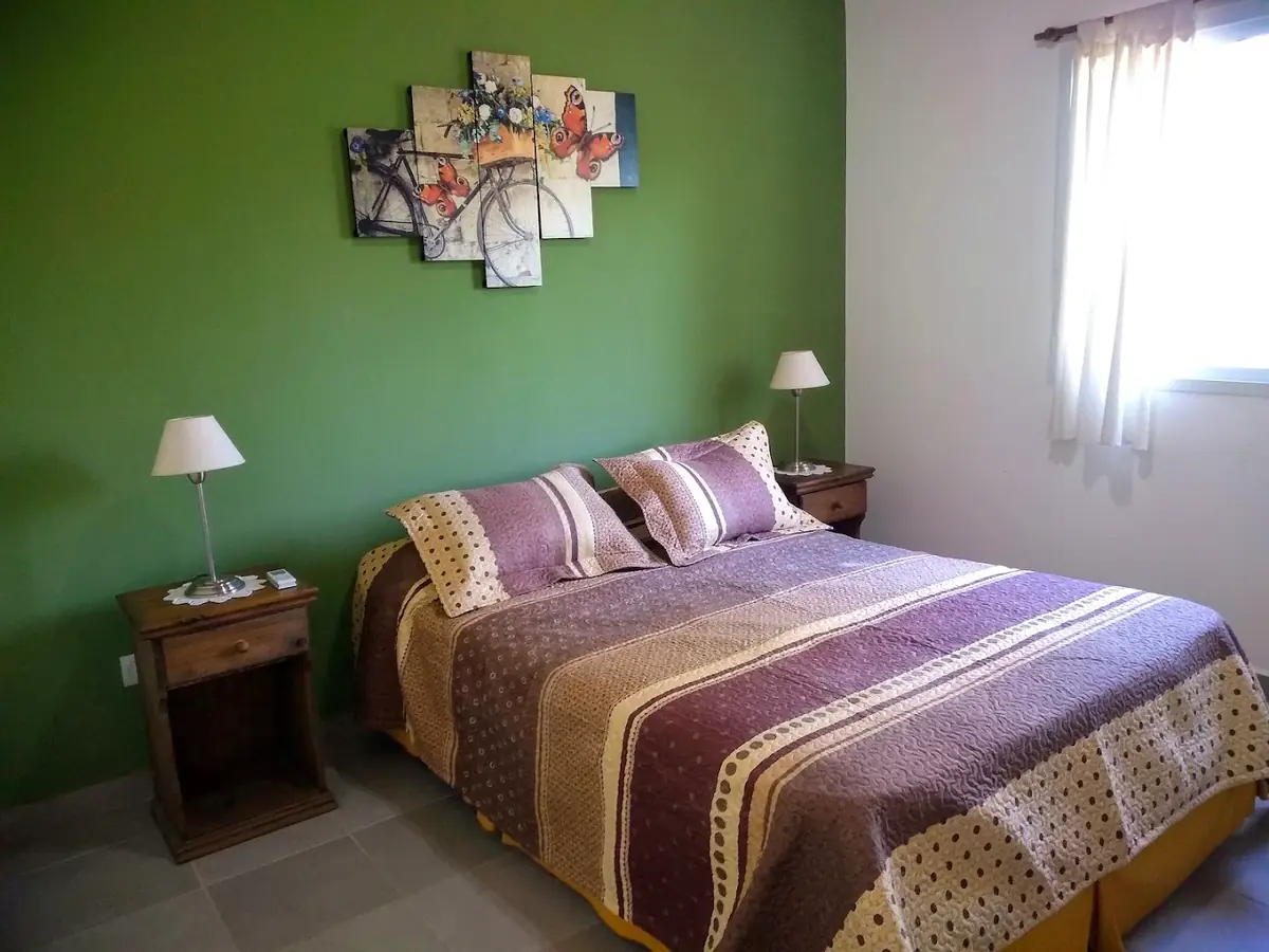 El dormitorio: cama matrimonial con acolchado a rayas, mesas de luz con veladores y pared verde.