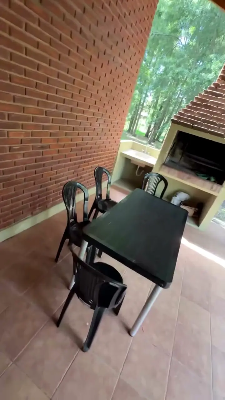 La galería techada con piso de cerámica, mesa y sillas, y la parrilla de ladrillo mirando al parque.