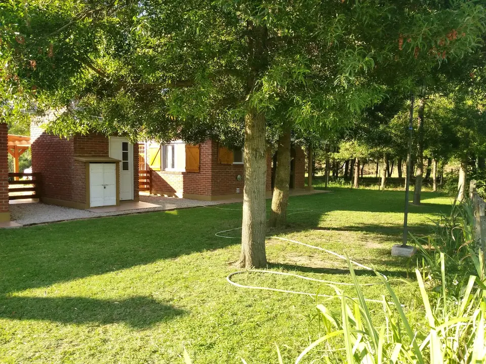 El parque de Chícharo: césped a la sombra de árboles grandes y una de las cabañas al fondo.