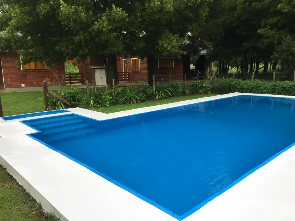 La pileta en forma de L con el agua celeste, el borde claro y el parque con árboles alrededor.