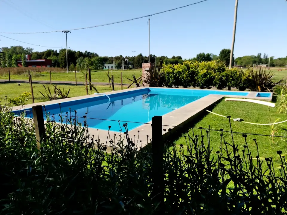 La pileta de Chícharo, de agua celeste, rodeada de pasto, plantas y un alambrado de campo, con árboles al fondo.