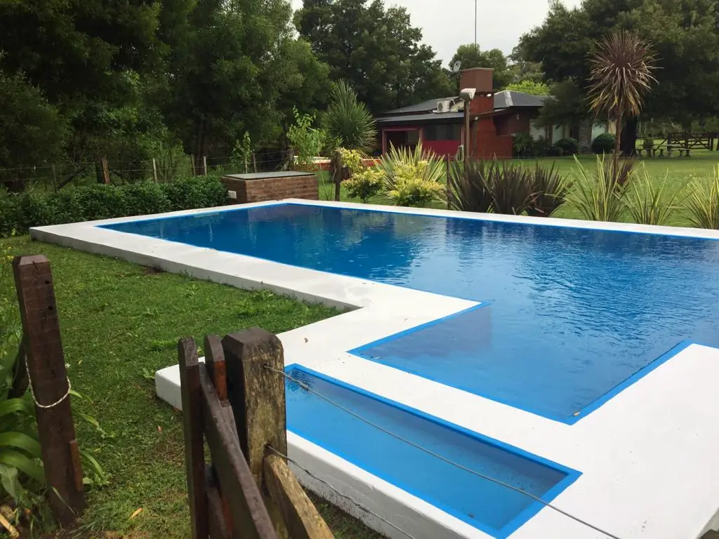 La pileta en forma de L, de agua celeste, con una tranquera de madera y el parque detrás.